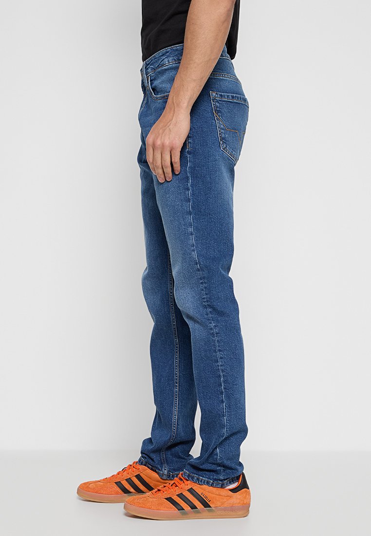 Blaue Jeans aus Denim mit slim fit, sichtbaren Nähten und einem Fünf-Taschen-Design. Kombiniert mit orangefarbenen und schwarzen Sneakers.