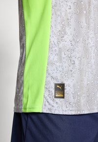 Szürke futballmez texturált, vízcsepp mintával; élénkzöld hálós betéttel és fekete-arany logócímkével az alján.