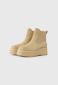 Beige semskede plattform ankelsko med ribbet elastiske sidepaneler og løkker, vist på en hvit bakgrunn.