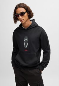 Svart hettegenser med et grafisk motiv av en åpen munn med spisse tenner, rødt "HUGO"-tekst og kengurulomme. Modellen har på seg svarte solbriller.