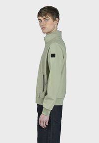 Lys grøn jakke med høj krave, sidelomme med lynlås, elastiske cuffe og et sort logo-patch på venstre ærme.