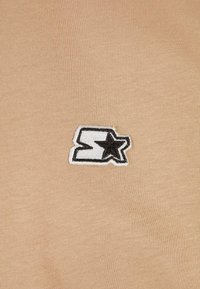 Beige színű, textúrázott megjelenésű pamut szöveten egy fekete-fehér hímzett "S" és csillag logó található.