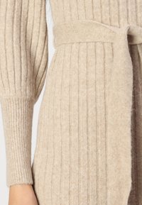 Pull en maille côtelée beige avec une taille ceinturée et des manches ajustées au fini texturé, mettant en valeur son matériau doux et confortable.