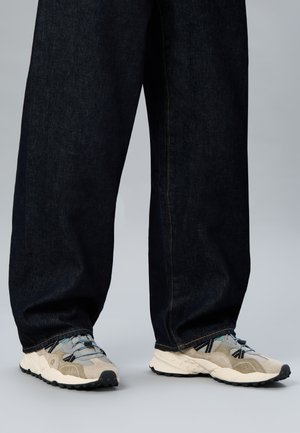 Pantaloni di jeans scuri a gamba larga indossati con sneakers chunky grigio chiaro e beige su sfondo grigio.