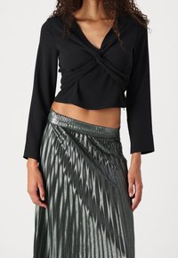 Blusa nera corta con design frontale annodato e maniche lunghe, abbinata a una gonna plissettata verde metallizzato con una texture lucida.