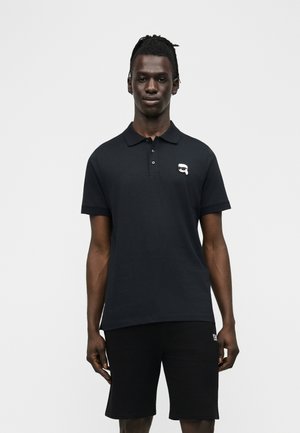 Černý mužský model s dredy, oblečený v černém polo tričku s krátkými rukávy a černých kraťasech, na světlém pozadí.