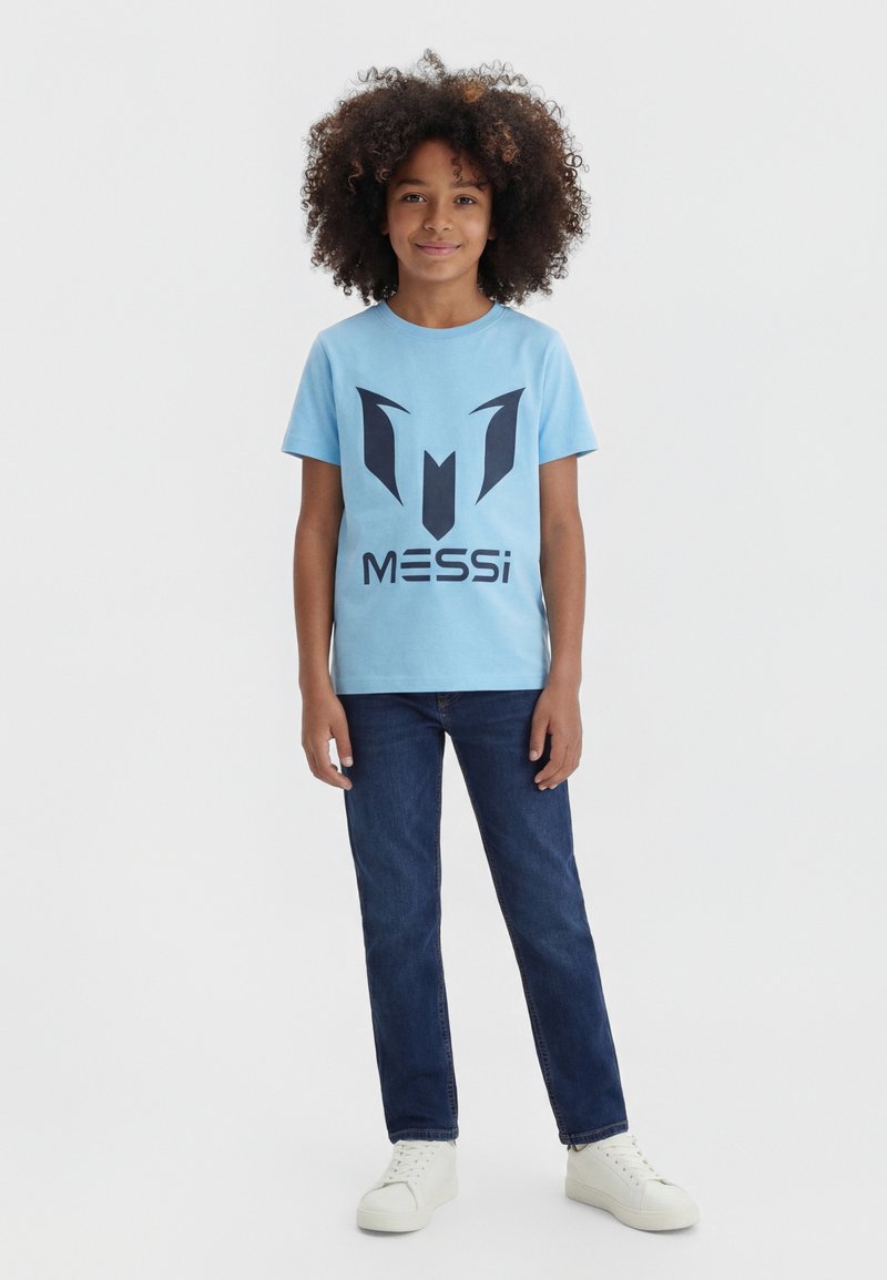 Kind met krullend haar dat een lichtblauw Messi T-shirt, donkerblauwe spijkerbroek en witte sneakers draagt, staand tegen een effen achtergrond.