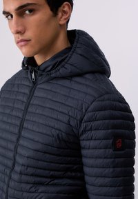 Jeune homme portant une veste matelassée à capuche bleu foncé avec une fermeture éclair à l'avant et un petit patch logo sur la manche gauche, faisant face à gauche.