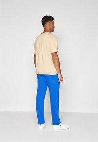 Beige t-skjorte med korte ermer, kombinert med blå bukser, med et enkelt design, myk tekstur, og hvite joggesko i bakgrunnen.