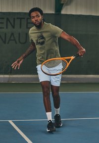 Green t-shirt with a black print, light blue shorts, and black athletic shoes. Holding an orange tennis racket on a blue court.