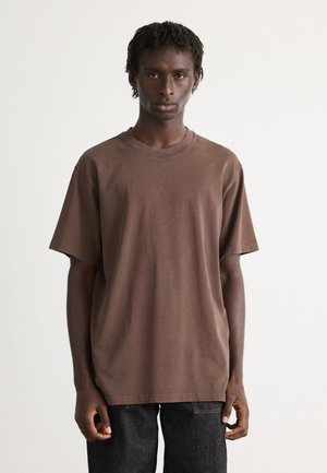 Young man with short dreadlocks wearing a plain brown oversized t-shirt and black jeans, standing against a white background.