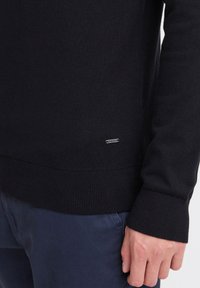 Svart strikkegenser med ribbestrikket nederkant og mansjetter, med en liten merkeflaske i metall nær bunnen. Myk tekstur og tettsittende design.