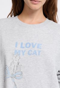 Primo piano di un felpa grigia con testo azzurro chiaro "ADORE IL MIO GATTO" e un disegno di un gatto seduto su un fiocco di nastro.