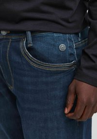 Mørkeblå denimjeans med synlig søm, en frontlomme og knappelukkelse. Nærbilde viser en hånd som hviler nær stoffet.