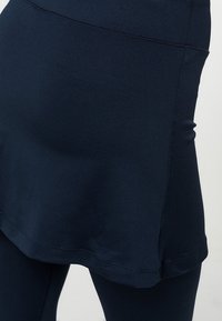 Sukně v námořnické modři s hladkou texturou, která má zakřivený lem a švy, doplněná o přiléhavé legíny pod ní.