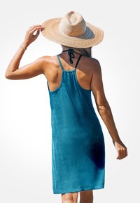 Türkisfarbnes Velours-Top-Kleid mit Racerback-Design. Weiche Textur, knielang, und kombiniert mit einem Strohhut, der von einer Hand gehalten wird.