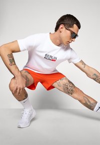 Man with tattoos wearing sunglasses, white "Berlin Run" t-shirt and orange shorts stretching with one leg bent and the other extended.