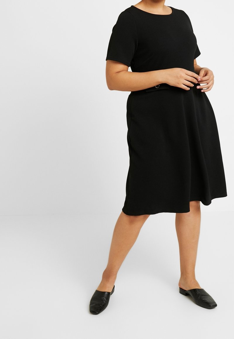 Femme en robe noire à manches courtes et longueur genoux avec détail ceinture latérale, portant des chaussures noires sans lacets, debout sur un fond blanc uni.