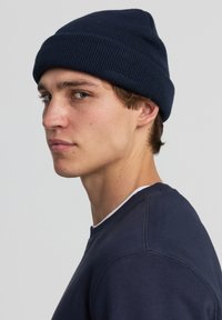 Giovane uomo che indossa un beanie blu navy e una felpa, mentre guarda oltre la spalla contro uno sfondo chiaro e semplice.
