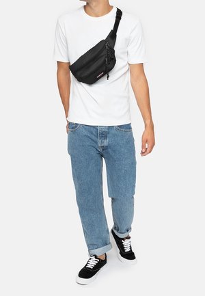 Zwarte nylon heupzak gedragen over een wit shirt, met een ritscompartiment en verstelbare band, gecombineerd met losse blauwe jeans en zwarte sneakers.