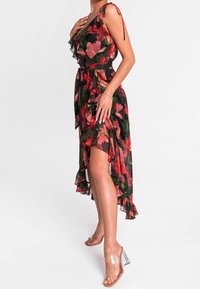 Robe florale avec une base noire ornée de motifs rouges et verts, des détails volantés, une taille ceinturée et un ourlet asymétrique. Accessoirisée avec des talons transparents.