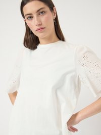 Mlada ženska z ravnimi rjavimi lasmi nosi belo bluzo z vezenimi nabranimi rokavi in črnimi visečimi uhančki, pozira pred belim ozadjem.