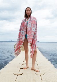 Young woman standing barefoot on floating pier by sea, wrapped in pink and blue patterned towel, cloudy sky overhead.
