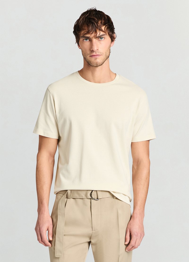 T-shirt beige à manches courtes en tissu texturé avec un col rond, associé à un pantalon crème avec passants de ceinture et une coupe slim.