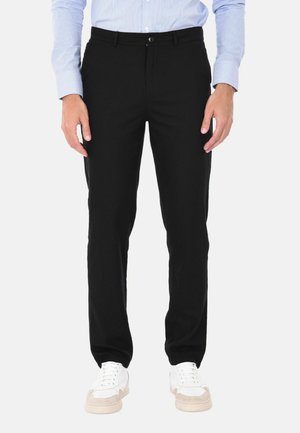 Uomo che indossa pantaloni neri a taglio slim, camicia a righe azzurra chiara e scarpe da ginnastica bianche, in piedi davanti a uno sfondo semplice.