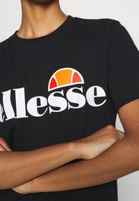 Camiseta de algodón negra con un gran logo blanco de "Ellesse" y un gráfico de sol colorido en naranja y rojo encima del logo. Mangas cortas.