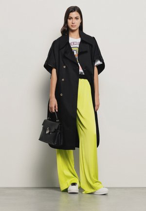 Trench coat nero doppiopetto con maniche ampie, abbinato a pantaloni larghi giallo neon e sneakers bianche, mentre tiene una borsa nera.