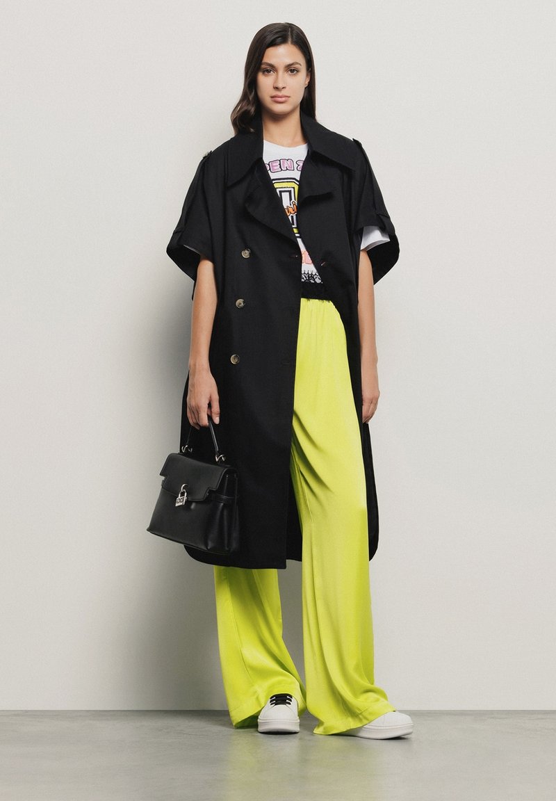 Trench coat nero doppiopetto con maniche ampie, abbinato a pantaloni larghi giallo neon e sneakers bianche, mentre tiene una borsa nera.