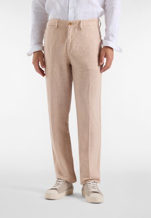 Man wearing beige linen pants, white long-sleeve button-up shirt, and beige sneakers standing against plain background.