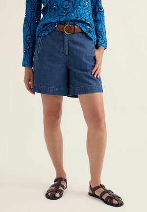 Nő kék virágmintás hosszú ujjú ingben, magas derekú farmer rövidnadrágban barna övvel és sötétbarna pántos szandálban.