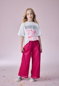 T-shirt bianco con testo grafico rosa e fiocchi, abbinato a pantaloni a gamba larga lucidi con paillettes rosa. Modello indossa sneaker bianche.