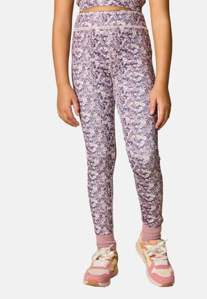 Kind trägt violette Leggings mit Blumenmuster, pinke Socken und pink-weiße Sneaker und steht mit locker an den Seiten hängenden Armen.