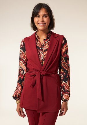 Bordeaux mouwloze vest met een strik in de taille, gelaagd over een zwart en rode blouse met lange mouwen met een paisley ontwerp.