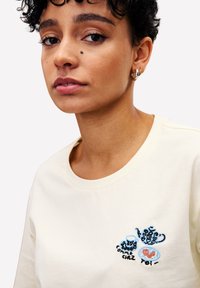 Jongere met kort krullend haar draagt een crèmekleurig shirt met geborduurde theepot, kopje en schotel met de Franse tekst "comme chez toi".