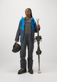 Laskijapuku, joka sisältää sinisen ja harmaan vedenpitävän takin, harmaan aluskerroksen, harmaat hiihdonhousut, mustat hiihtokengät, hiihtokypärän ja sukset.