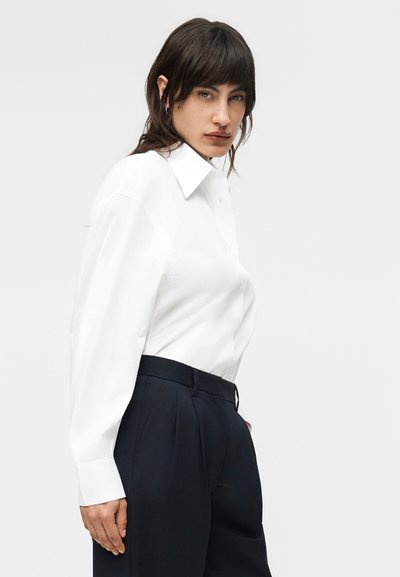 Tumejuustega naine, kes kannab valget kaelusega pikkade varrukatega särki ja kõrge vöökohaga musti pükse, vaadates küljele ühevärvilise tausta ees.