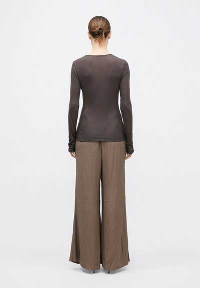 Nő kontyba fogott hajjal, szűk, hosszú ujjú sötétbarna felsőt és bőszárú világosbarna nadrágot viselve, egyszínű fehér háttér előtt áll.