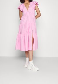 Różowa bawełniana sukienka o wielowarstwowym kroju, z krótkimi falbaniastymi rękawami i szczeliną z przodu. W zestawieniu z białymi, masywnymi trampkami.