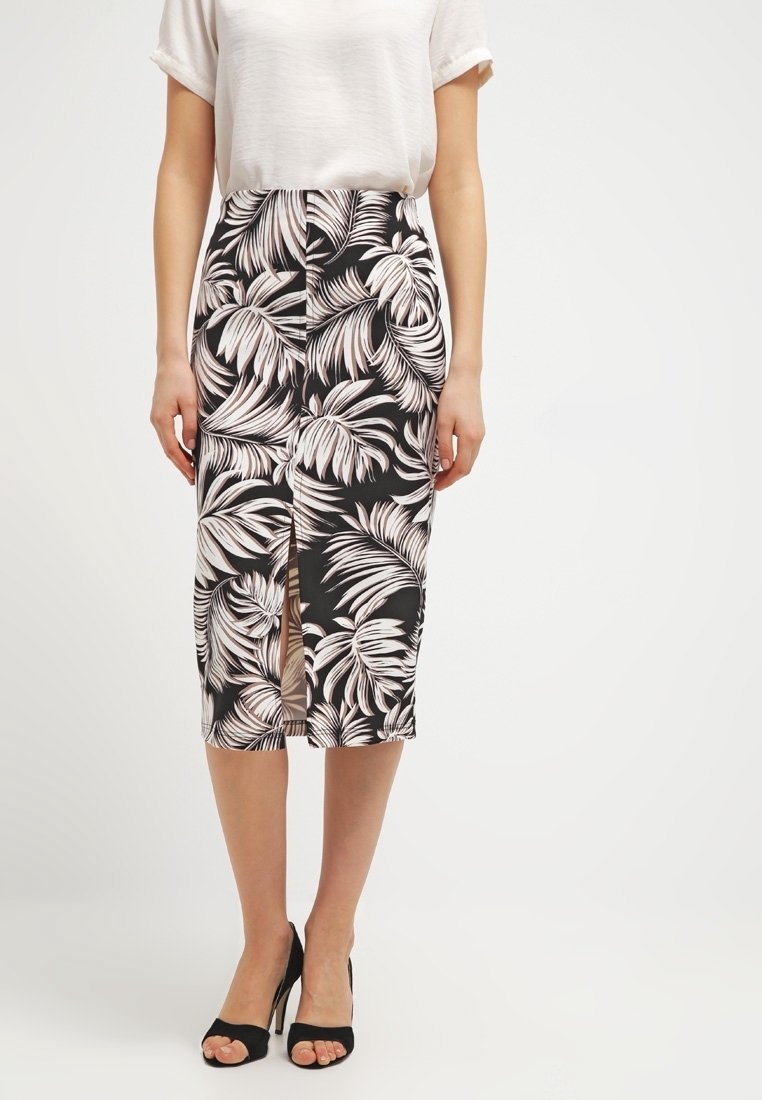 Femme portant une jupe midi à imprimé feuilles tropicales noir et blanc avec une fente devant, un top blanc à manches courtes et des talons ouverts noirs.
