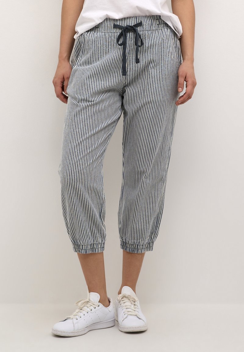 Pantalons courts rayés en bleu et blanc, avec une taille élastique dotée d'un cordon de serrage et des poignets élastiques. Associés à des baskets blanches.