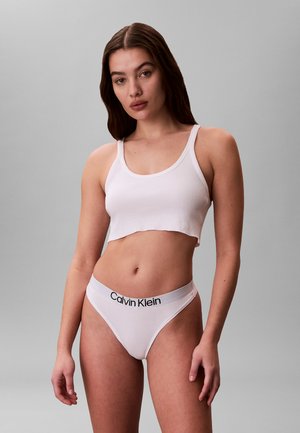 Ung kvinde med langt brunt hår iført en hvid kort tanktop og hvide Calvin Klein undertøj står mod en ensartet grå baggrund.