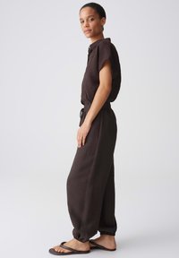 Woman standing sideways wearing dark brown short-sleeve button-up shirt, matching drawstring pants, and dark brown sandals against a white background.