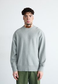 Sudadera gris con cuello redondo y mangas largas, que presenta un dobladillo y puños acanalados. Combinada con pantalones verdes, sobre un fondo claro.