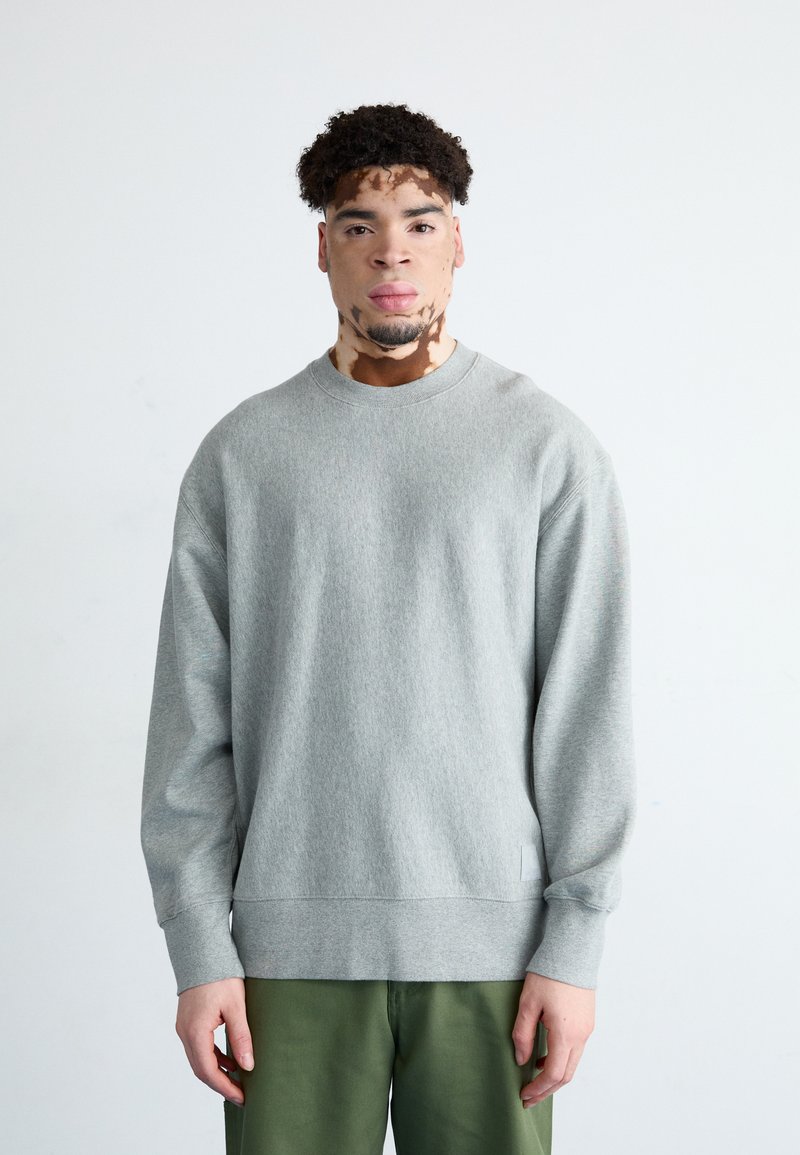 Sudadera gris con cuello redondo y mangas largas, que presenta un dobladillo y puños acanalados. Combinada con pantalones verdes, sobre un fondo claro.