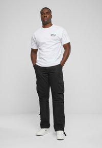 White short-sleeve t-shirt with black graphic on the left chest, paired with black cargo pants featuring side pockets and white sneakers.