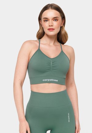 Green bralette with a scoop neckline, ruched detailing, and logo at the hem. Fabric is smooth and stretchy, paired with high-waisted leggings.
